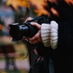 A young woman captures a moment outdoors with a camera amidst autumn foliage.
